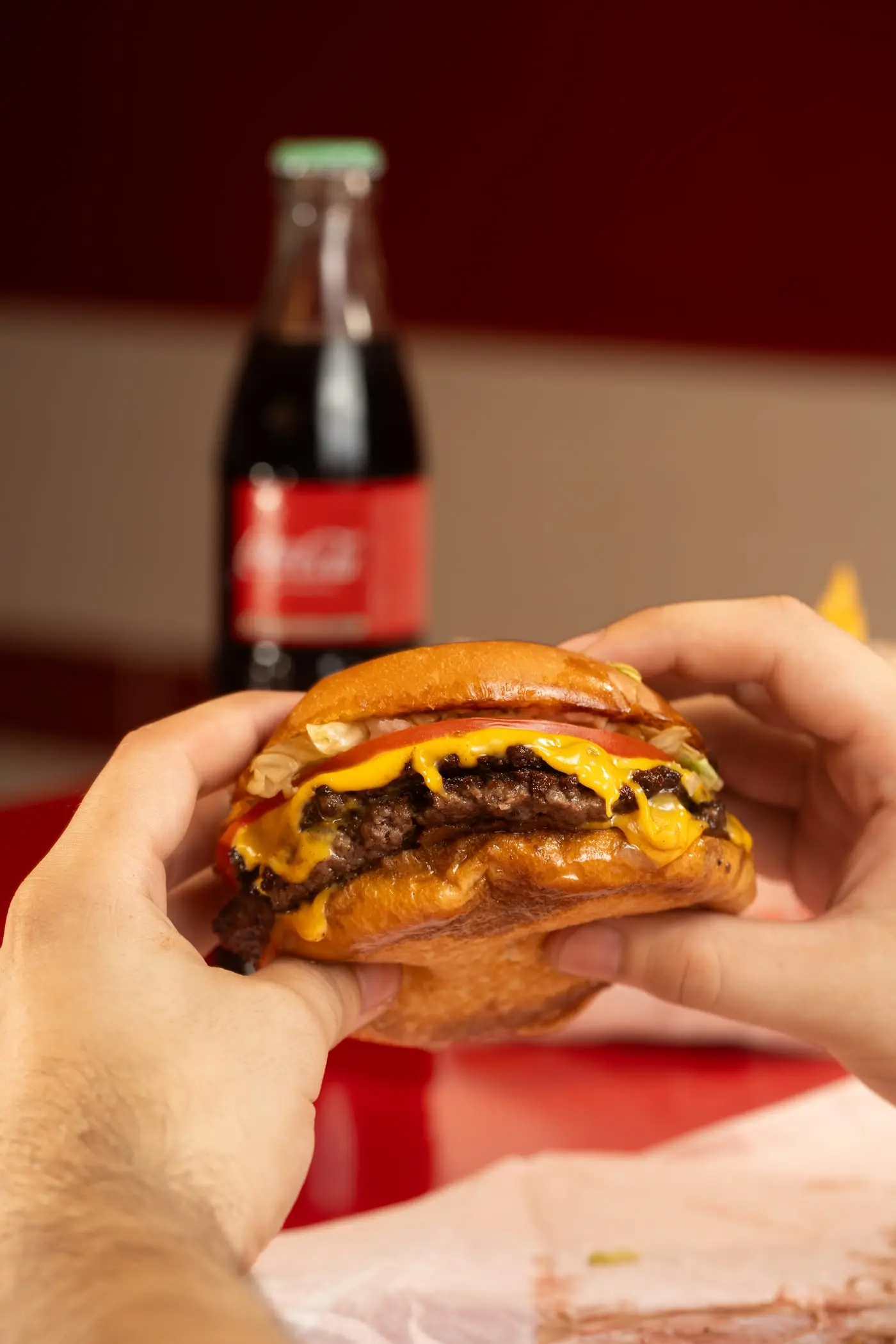 Manos sosteniendo una hamburguesa smash frente a una Coca-Cola de vidrio
