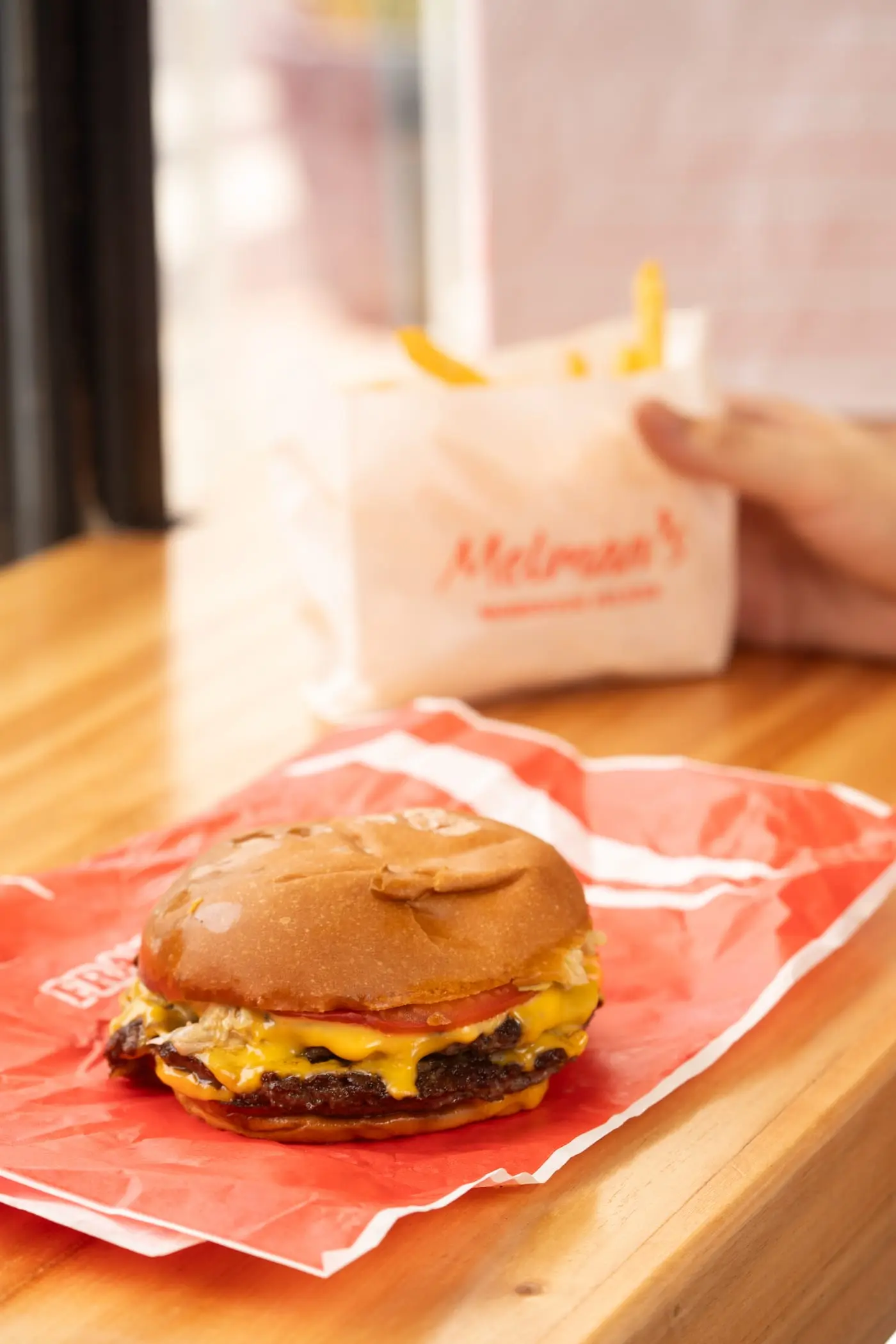 Hamburguesa doble con queso sobre su papel rojo, con una bolsa de papas detrás